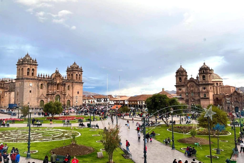 A wide shot of the Plaza de Armas in Cusco, showing the cathedral buildings, crowds of people, and beautifully arranged gardens in the foreground. The sky is partly cloudy, with the Andean mountains visible in the background.