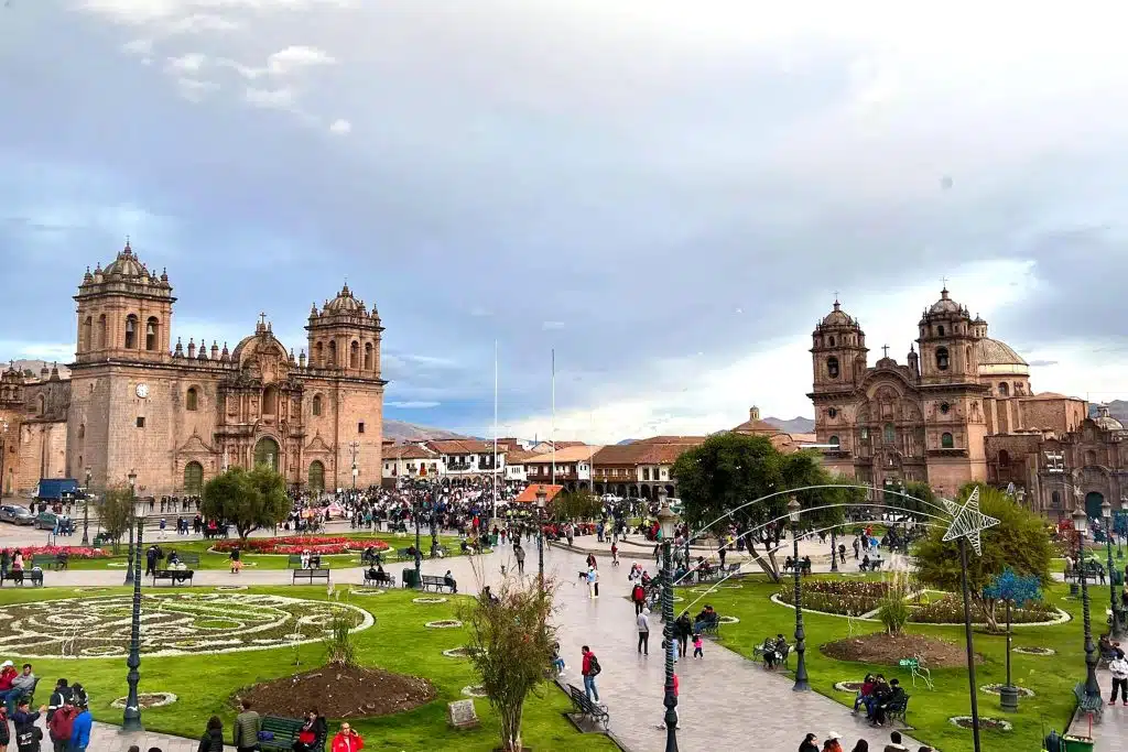 A wide shot of the Plaza de Armas in Cusco, showing the cathedral buildings, crowds of people, and beautifully arranged gardens in the foreground. The sky is partly cloudy, with the Andean mountains visible in the background.