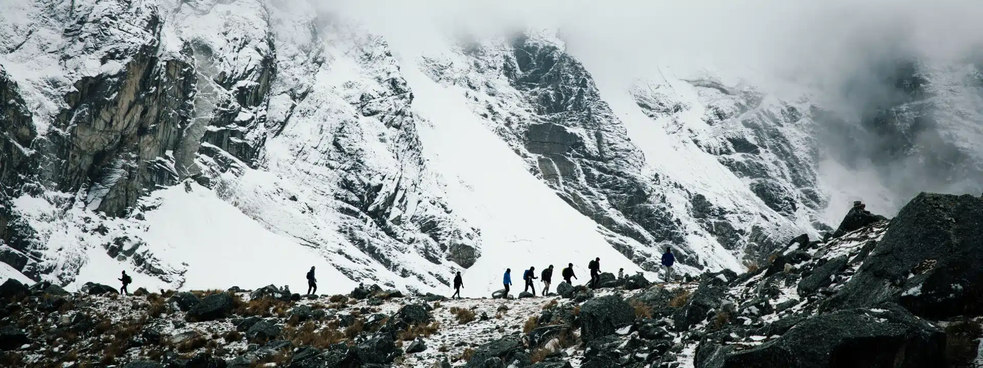 Hikers crossing a rocky, snow-covered trail beneath the towering peaks of Salkantay Mountain in the Peruvian Andes.