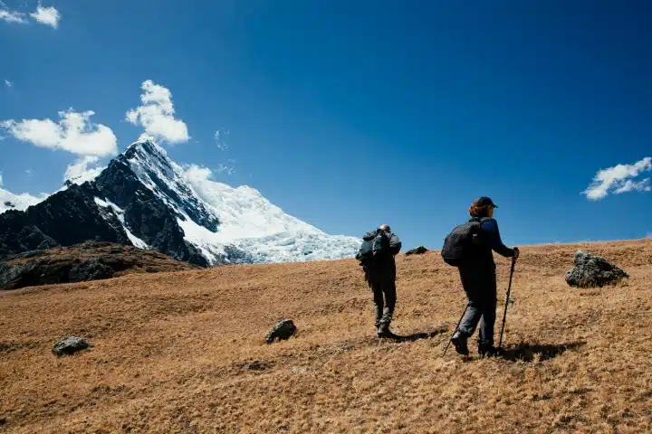 Salkantay Mountain: History, Meaning and Spiritual Significance 1 Two hikers walking across a golden mountain landscape with a snow-capped peak along the Salkantay Trek in Peru.